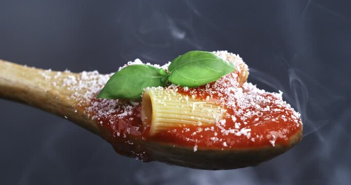 Super slow motion macro of fresh green basil leaf is falling on traditional bio tomato sauce with pasta with grated parmesan cheese in wooden spoon isolated on soft background at 1000 fps.