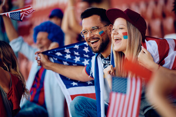 Happy couple of sports fans wrapped in USA national flag watching game at stadium.