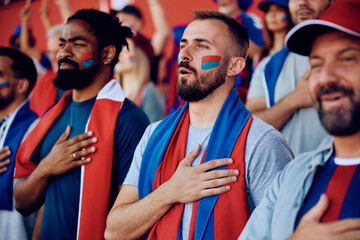 Group of sports fans with hand on heart singing national anthem during championship at stadium. - obrazy, fototapety, plakaty