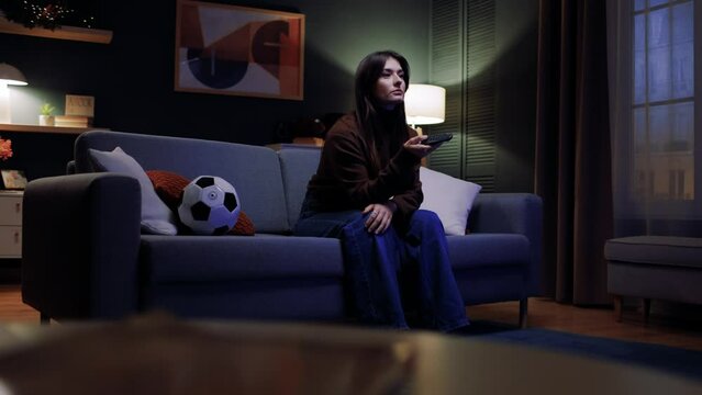 Female sitting at couch with cushions, smiling while watching TV set. Excited joyful young woman watching football match, cheering up for favorite team, enjoying after scored goal.