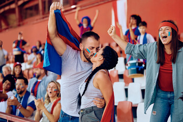 Couple of sports fans kissing while celebrating during match at stadium.