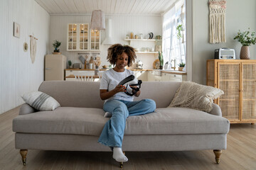 Millennial African American girl with curly hair sitting on couch testing new VR headset, using virtual reality helmet to view photos and videos in 3D. Young woman examining AR glasses at home