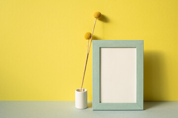 Photo frame with vase of dry flower on gray shelf. yellow wall background