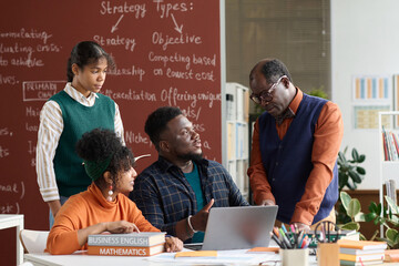 Portrait of senior African American teacher talking to group of students sitting at table with...