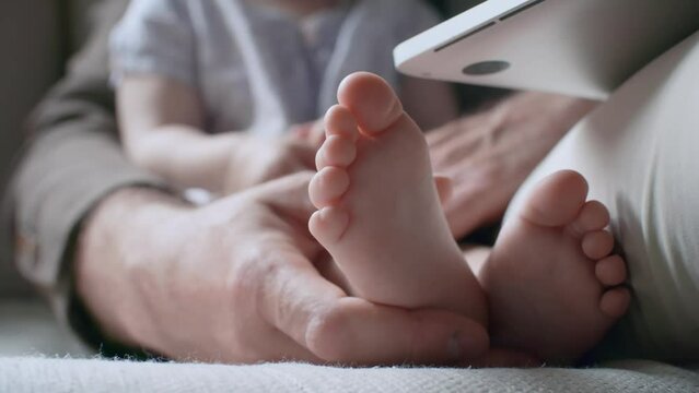 Handheld close up shot of hands of unrecognizable father holding little feet of toddler girl sitting beside him and watching cartoons on laptopHandheld close up shot of hands of unrecognizable father 