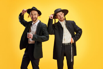 Jewish men in traditional suit and hats posing holds glass of alcohol drinks against vivid yellow background. Purim, business, festival, holiday, celebration Pesach or Passover, Judaism concept.