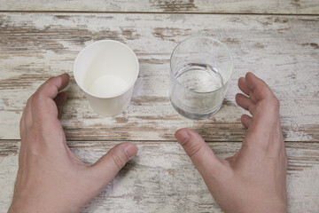 A glass cup and a paper cup stand side by side on a wooden background with hands reaching for them. Glass or disposable tableware, choice and choice problems