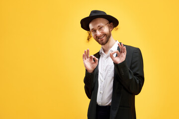 Portrait of smiling man in suit and black hat making positive hand gesture against sunny yellow...