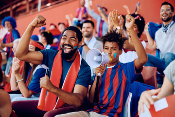 Cheerful black father and son cheering while watching sports match at stadium.