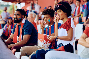 Black family eating popcorn while watching sports game at stadium.