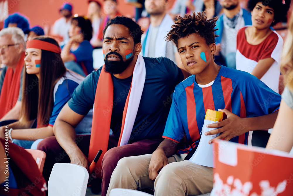 Wall mural Black sports fan and his son watching the game from the stadium stands.