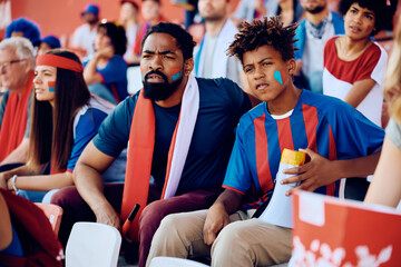 Black sports fan and his son watching the game from the stadium stands.