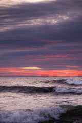 A pattern of three waves making their way to the beach and crashing on the shallow water. Each wave has a whitecap. The ocean is beneath a cloud filled sky lit by the rising sun with pink and orange.