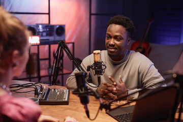 Young African American man carefully listening to unrecognizable woman sitting at desk with studio equipment int podcast room with neon light