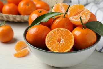 Delicious tangerines with green leaves in bowl on white wooden table, closeup