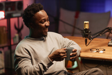 Cheerful young African American male blogger drinking coffee during live podcast in recording studio