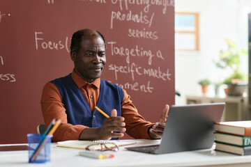 Portrait of senior Black college professor sitting at desk and using computer, copy space