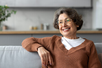 Relaxed senior woman with grey hair in glasses sitting on couch and looking aside, resting in...