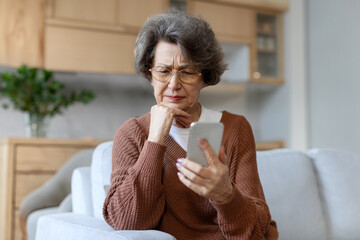 Thoughtful elderly woman in glasses looking at phone screen with serious and sad expression,...