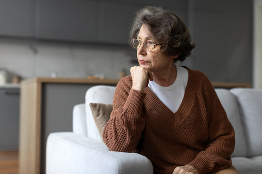 Depressed senior woman sitting on sofa, gazing out of window with thoughtful expression, capturing moment of solitude in her quiet space