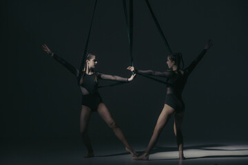 Fototapeta na wymiar Portrait of two female acrobats in studio isolated on black background. Girls aerial dancers performing element with ropes.