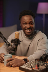 Portrait of happy Black man looking at camera holding cup sitting at table with audio equipment in podcast studio