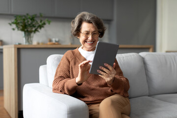 Joyful senior woman, smiling brightly and using digital tablet while sitting on cozy sofa at home, creating warm and modern lifestyle scene