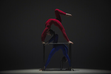Portrait of two female acrobats isolated on black background. Girls aerial dancers in blue red suits showing element on a cube.