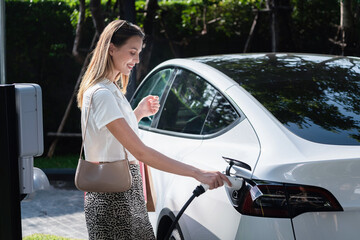 Young woman travel with EV electric car charging in green sustainable city outdoor garden in...