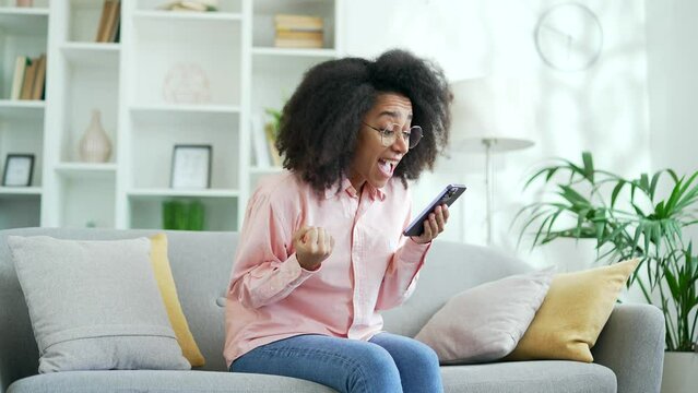 Excited happy young african american female celebrating success by reading great news on smartphone sitting on sofa in living room at home. Smiling black woman satisfied with positive message on phone
