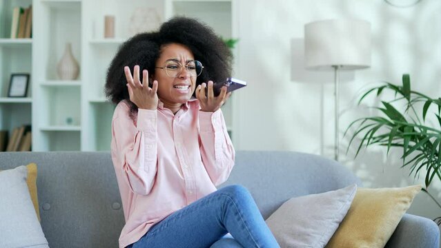 Angry young african american female talking arguing on the mobile phone while sitting on sofa in living room at home. Frustrated excited black woman says screams about problems using a smartphone