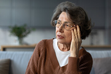 Upset senior woman in glasses sitting on sofa at home, touching her temples and looking aside with...