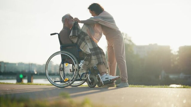 Wife helps husband to sit in wheelchair. Woman covers elderly man with blanket, walking together in park. Caring female help male with special needs, physical disability, lifelong love concept.