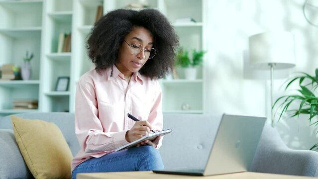 Young african american female student listens to online e-learning, video course using a laptop sitting at home. Black woman at a remote learning seminar or training writes in notebook talks to tutor