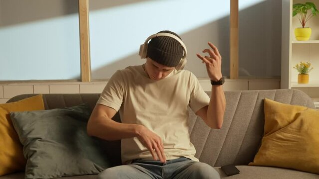 Portrait of young person in the room sitting on the couch. Man in hat on the sofa in headphones listening music playing on air guitar.