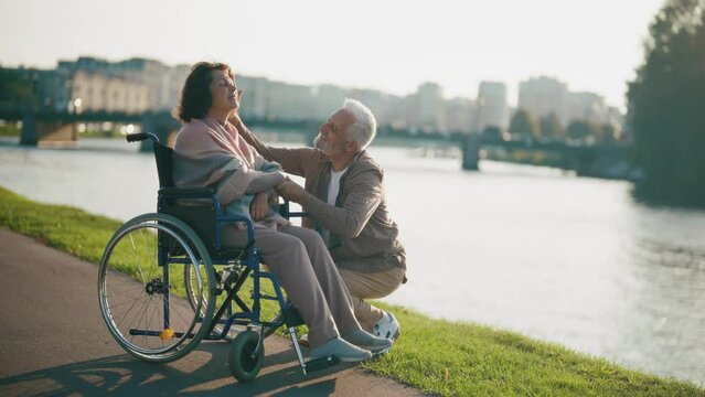 Husband walking with wife in wheelchair. Senior family couple spending time in city park. Man hugging female showing love care, they smiling. Living with physical disability, together love concept.