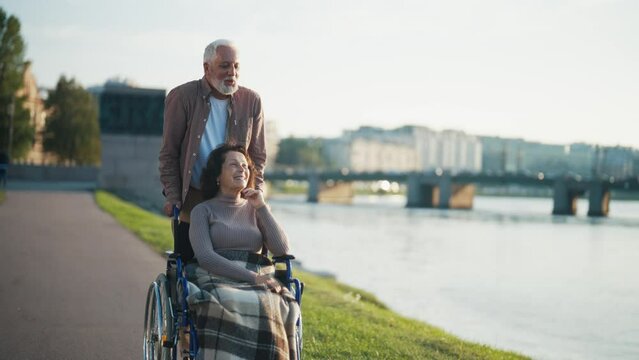 Elderly husband walking with wife in wheelchair. Senior family couple spending time in city park near river. Man caring woman showing love, smiling communication. Living with physical disability.