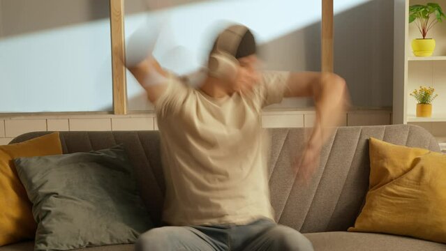 Portrait of young person in the room sitting on the couch. Man in hat on the sofa in headphones jams to music dances imitates sounds.