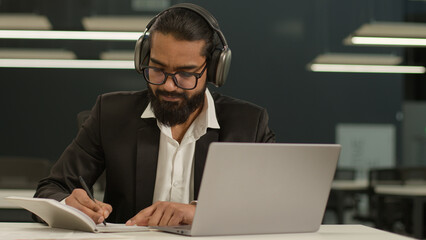Focused Arabian male Indian businessman wear headphones ethnic business man studying working in office with laptop writing notes write in notebook video conference call wearing wireless headphones - obrazy, fototapety, plakaty