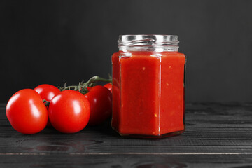 Delicious ketchup in jar and tomatoes on black wooden table. Tomato sauce