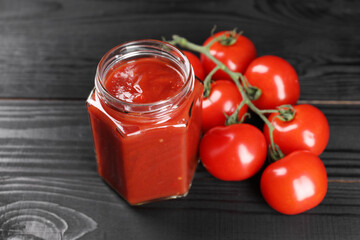 Delicious ketchup in jar and tomatoes on black wooden table, closeup. Tomato sauce