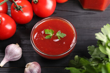 Delicious ketchup in bowl, garlic, parsley and tomatoes on black wooden table, closeup. Tomato sauce