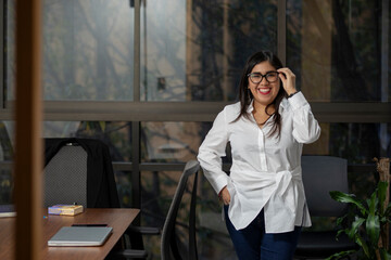 Portrait of a Mexican woman standing in her office smiling effusively while looking at the camera and holding her glasses.
