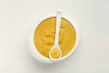 Spoon and bowl of tasty mustard sauce on white wooden table, top view