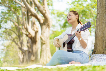 Asian beautiful woman sits under the tree at the park and enjoy playing an acoustic guitar. Woman...