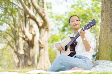 Asian beautiful woman sits under the tree at the park and enjoy playing an acoustic guitar. Woman...