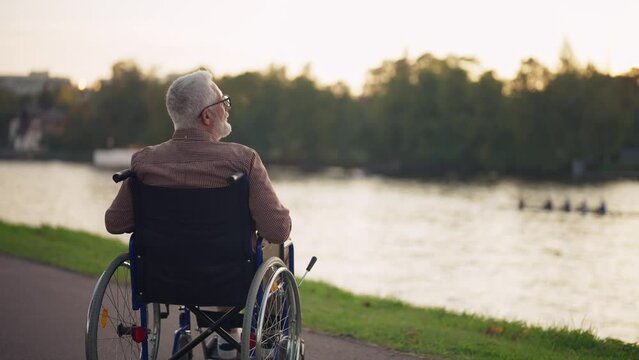 Man in wheelchair walking in city park. Back view senior male breathing fresh air near pond admiring enjoying nature in evening. Live with physical disability, special needs people in old age concept.