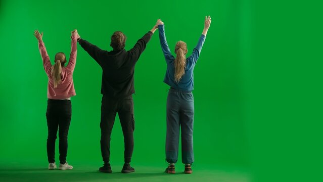 Back view of a team of three hikers raising their hands in the air, making a gesture of victory. A group of people rejoicing and cheering in a studio on a green screen. Travel concept. - Powered by Adobe