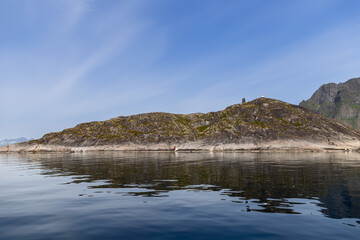 A serene Scandinavian fjord with a lone lighthouse atop a moss-covered rocky island, its reflection on the calm sea water under a clear blue sky. Lofoten, Norway