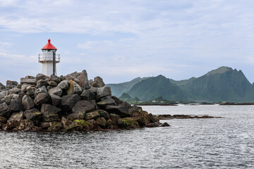 A lighthouse with a vivid red cap sits atop a stone jetty at Andenes in the Lofoten Islands, Norway, with lush mountains and a subdued sky framing the seascape - obrazy, fototapety, plakaty
